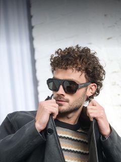 Ein Mann mit Sonnenbrille, schwarzer Lederjacke und gestreiftem Pullover steht vor einer weißen Backsteinwand und rückt mit einer Hand den Kragen seiner Jacke zurecht.