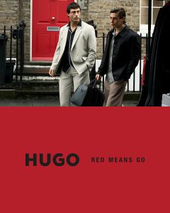 Zwei Models in schwarzen Anzügen posieren selbstbewusst vor einem schlichten hellen Hintergrund. Das HUGO Logo erscheint in fetten weißen Buchstaben in der unteren linken Ecke.