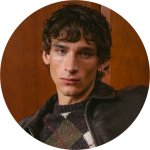Mann mit lockigem braunem Haar und gestutztem Bart, der einen hellen Blazer und ein weißes Hemd trägt, steht vor einem weißen Hintergrund und lächelt sanft.