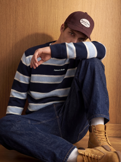Ein junger Mann in Jeansjacke und Jeans kniet nieder und schaut zur Seite. Er trägt weiße Turnschuhe und hat eine sichtbare Tätowierung auf seinem Unterarm. Der Hintergrund ist einfarbig hellgrau.