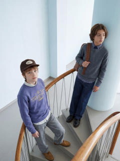 Zwei Jungen in Jeans und Jeanshemden posieren zusammen. Der eine sitzt auf einem Hocker, hat die Hände in den Schoß gelegt und einen Schal um den Hals, während der andere mit dem Arm auf dem Hocker steht und eine Mütze trägt.