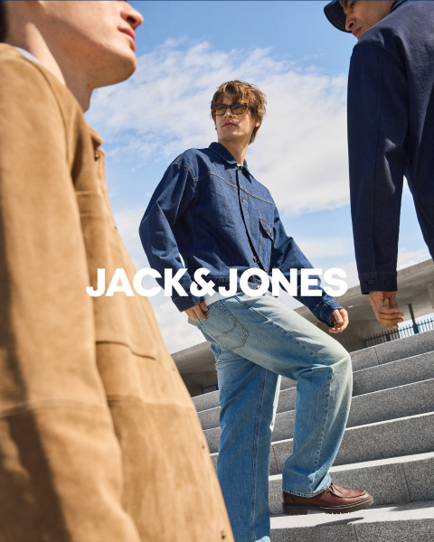 Zwei Männer in Jeans-Outfits sitzen vor einem schlichten Hintergrund. Der vordere Mann hält sich die Hand auf den Mund, während der andere hinter ihm sitzt. In der Mitte steht der Text "JACK & JONES".