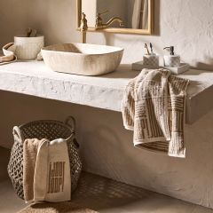 Minimalistisches Badezimmer mit rundem Spiegel, wandmontiertem Wasserhahn und rechteckigem Waschbecken. Regale halten Dekorationsgegenstände und Handtücher in Erdtönen. Sanftes Licht wirft sanfte Schatten auf die strukturierte Wand und schafft eine ruhige Atmosphäre.