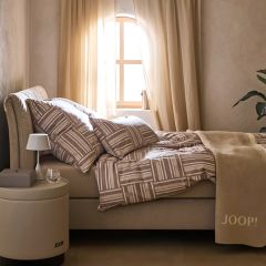 Ein gemütliches Schlafzimmer mit einem beigefarbenen Bett mit gemusterter Bettwäsche. An der Wand hängt ein runder Spiegel. Sanftes Licht beleuchtet einen Teil des Bettes. Ein kleiner Stapel Bücher und Dekorationsgegenstände liegen in der Nähe auf dem Boden.