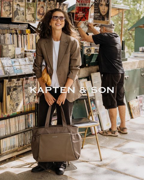 Eine Frau und ein Mann lächeln sich im Freien an. Die Frau trägt eine grüne Mütze und einen beigen Mantel und hält eine schwarze Tasche in der Hand. Der Mann trägt eine braune Jacke und eine cremefarbene Tasche. In der Mitte steht „Kapten & Son“.