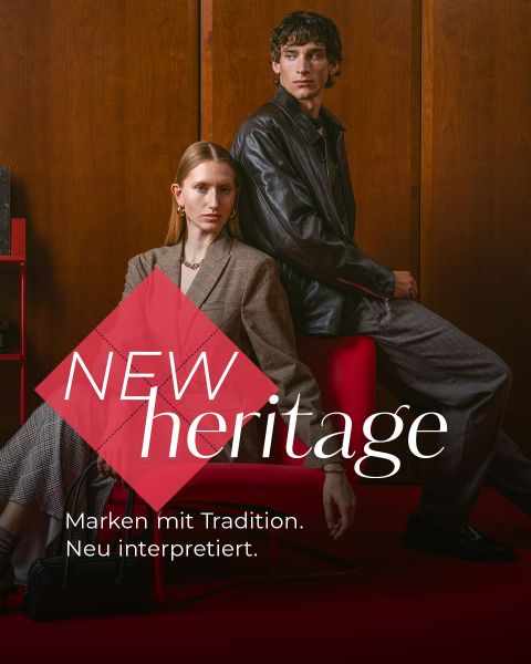Zwei Models in klassischen Outfits posieren in einem mit Holz vertäfelten Raum mit roten Stühlen. Der Text lautet: „NEW heritage. Marken mit Tradition. Zeitlose Looks.“.