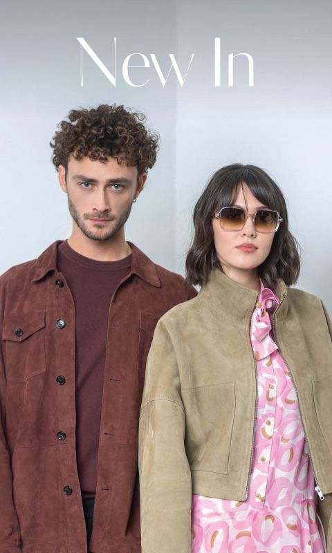 Ein Mann in einer braunen Jacke und eine Frau mit Sonnenbrille und hellbrauner Jacke über einem rosa gemusterten Kleid stehen nebeneinander vor einem schlichten hellen Hintergrund.