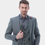 Mann in einem grauen dreiteiligen Anzug mit gemustertem Hemd und roter Reversnadel, der selbstbewusst vor einem schlichten hellen Hintergrund posiert.