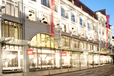 Ein hell erleuchtetes, elegantes Kaufhaus mit großen Glasfenstern und einem Glasvordach, fotografiert in der Abenddämmerung auf einer städtischen Straße.