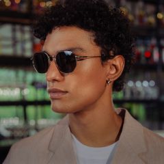 Eine Person mit kurzen lockigen Haaren, die eine Sonnenbrille, einen beigen Blazer und ein weißes Hemd trägt, steht in einem Innenraum mit Glasregalen im Hintergrund.