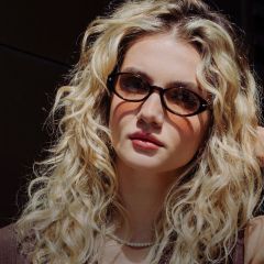 Junge Frau mit langem, lockigem blondem Haar, die eine Schildpattbrille, eine Perlenkette und ein braunes Oberteil trägt und in die Kamera schaut.