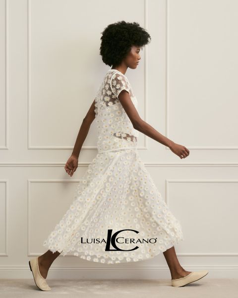 Eine Frau mit langen braunen Haaren trägt ein geblümtes Kleid und ein strukturiertes, rostfarbenes Pelzgewand über der Schulter. Sie blickt direkt in die Kamera. Unten in der Mitte ist der Text „Luisa Cerano“ zu sehen.