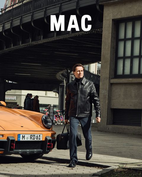 Eine Frau in einer Pelzjacke und weiten Hosen steht neben einem Mann in einer beigen Jacke und khakifarbenen Hosen. Beide posieren selbstbewusst im Freien vor einem steinernen Geländer unter blauem Himmel. Das Wort "MAC" erscheint am unteren Rand.