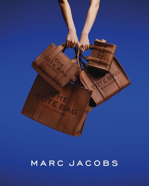 Eine Frau posiert im Freien mit einer braunen Marc-Jacobs-Tasche in der Hand, lehnt sich mit geneigtem Kopf zurück und trägt eine kastanienbraune Jacke und blaue Jeans. Auf dem Bild erscheinen die Worte "THE TOTE BAG MARC JACOBS" und "MARC JACOBS".