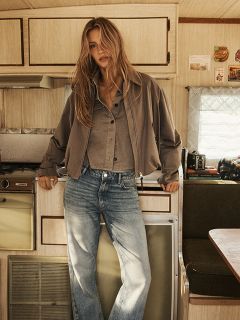 Eine Frau mit langem, gewelltem blondem Haar trägt einen braunen Kapuzenmantel mit schwarzem Gürtel, steht vor einer rustikalen Holzwand und blickt selbstbewusst in die Kamera.