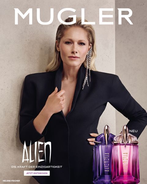 Eine Frau mit strahlend dunkler Haut und glattem schwarzen Haar posiert vor einem violetten Hintergrund. Vor ihr stehen zwei Alien-Parfümflakons von Mugler. Der Text lautet „MUGLER“ und „ALIEN EXTRA INTENSE THE REFILLABLE FRAGRANCES“.