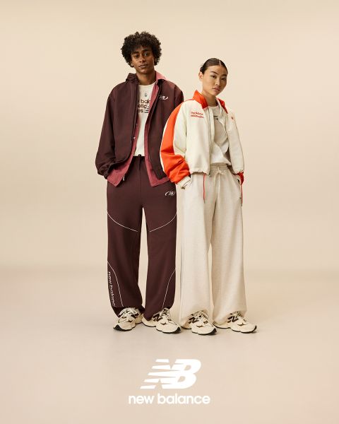 Ein Mann und eine Frau posieren vor Neonreklamen. Der Mann trägt einen beigen Trainingsanzug, die Frau einen Jeans-Overall. Beide tragen New Balance-Sneaker. Die Szene wird von verschiedenen Neonlichtern in Blau und Rot beleuchtet.