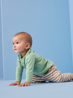 Ein lächelndes Baby liegt auf einer weißen Decke und trägt einen blauen Strickpullover, eine braune Cordhose mit Taschen und graue Socken. Das Baby sieht glücklich und entspannt aus und hat die Arme an den Ellbogen leicht angewinkelt.