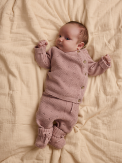 Ein Baby liegt auf einer weißen Decke und trägt einen hellrosa Strickpulli mit Petermännchenkragen und eine passende Hose sowie cremefarbene Socken.