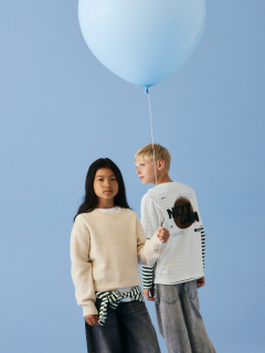 Ein kleines Kind mit blondem Haar steht lächelnd vor einem grünen Hintergrund und trägt einen braunen ärmellosen Pullover über einem geblümten Kleid, kniehohe Socken und braune Schuhe.