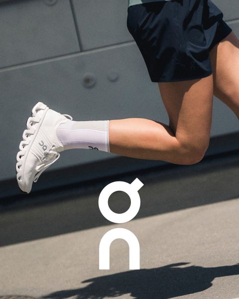Zwei Sneaker der Marke „On“ werden auf einem strukturierten Ast vor einem farbenfrohen Sonnenuntergangshimmel präsentiert. Ein Schuh ist schwarz mit grauen Akzenten und der andere ist weiß mit dunkleren Details. Darunter ist das „On“-Logo sichtbar.