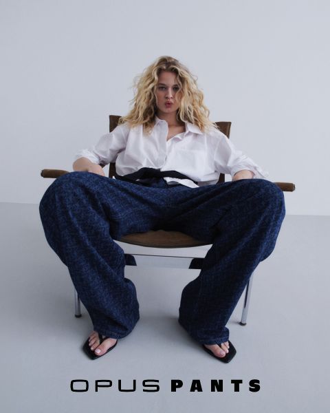 Person in lockerem weißen Hemd und Bluejeans, die Hände in die Hüften gestemmt. Unten steht der Text „Opus Pants“. Das Haar ist leicht vom Wind zerzaust, weißer Hintergrund.