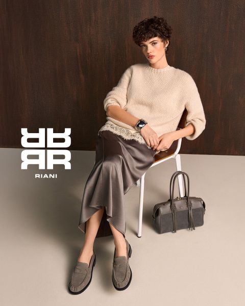 Zwei Frauen posieren stilvoll in einem Theater mit roten Sitzen. Die eine sitzt auf der Rückenlehne eines Sitzes und trägt ein gemustertes Kleid, einen schwarzen Mantel und Stiefel. Die andere steht in einem marineblauen Pullover und einem schwarzen Rock neben ihr. Über ihnen erscheint das Logo "RIANI".