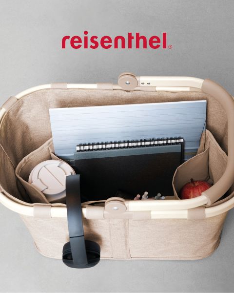 Eine Person mit langen roten Haaren, die ein hellblaues Hemd und weiße Hosen trägt, entspannt sich im Gras und hält eine gemusterte Tasche in der Hand. Sie steht draußen auf einem sonnigen Feld mit Blick auf die Hügel in der Ferne. Das Wort „Reisenthel“ ist in roter Schrift zu sehen.