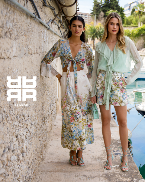 Zwei Frauen posieren stilvoll in einem Theater mit roten Sitzen. Die eine sitzt auf der Rückenlehne eines Sitzes und trägt ein gemustertes Kleid, einen schwarzen Mantel und Stiefel. Die andere steht in einem marineblauen Pullover und einem schwarzen Rock neben ihr. Über ihnen erscheint das Logo "RIANI".