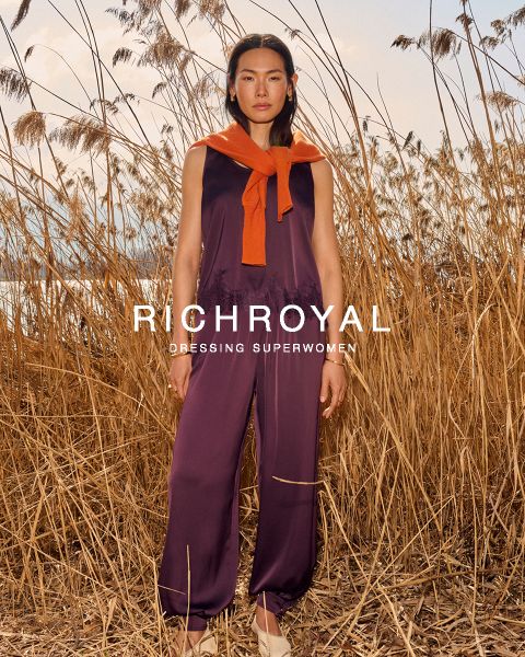 Eine Frau in hellblauem Hemd und grauer Hose hält sich eine Schneekugel vor ein Auge. Sie posiert in einem Raum mit Holzwänden und einem Skiplakat und trägt große Ohrringe. Der Text lautet "RICHROYAL DRESSING SUPERWOMEN".