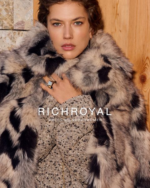 Eine Frau in hellblauem Hemd und grauer Hose hält sich eine Schneekugel vor ein Auge. Sie posiert in einem Raum mit Holzwänden und einem Skiplakat und trägt große Ohrringe. Der Text lautet "RICHROYAL DRESSING SUPERWOMEN".