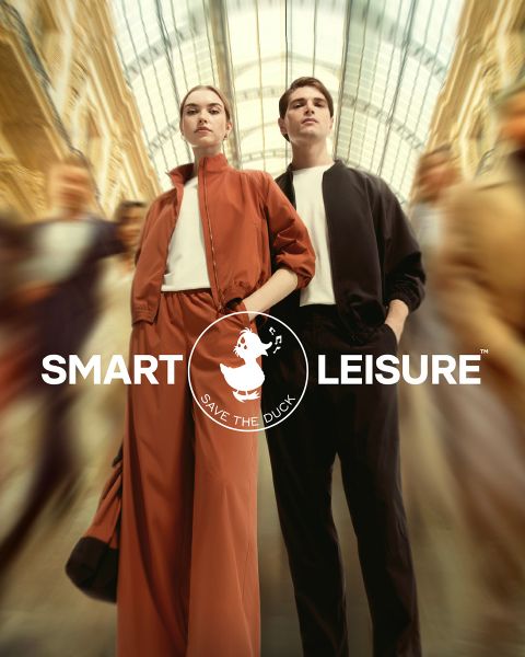 Ein Model läuft auf einem mit Blättern bedeckten Pfad. Es trägt eine orangefarbene Pelzjacke, einen passenden Rock und eine Mütze mit orangefarbenen Strumpfhosen und pfirsichfarbenen Socken. In der Ecke ist das Logo von „Save the Duck“ zu sehen.