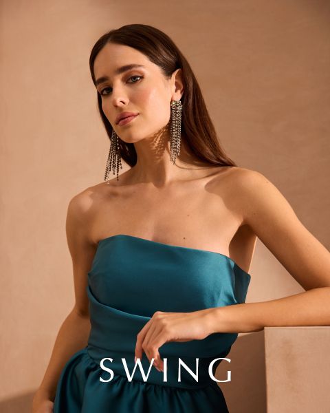 Eine Frau in einem seidigen, salbeigrünen Kleid sitzt auf einem cremefarbenen Stuhl vor einem floralen Hintergrund. Sie trägt goldene Absätze, Statement-Ohrringe und ein Armband. Das Wort "SWING" erscheint am unteren Rand des Bildes.