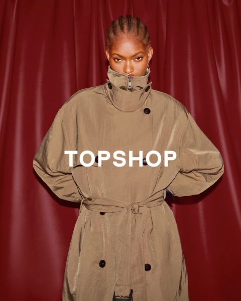 Eine Frau in einem übergroßen grünen Top, einer in der Taille gebundenen Jeansjacke, schwarzen Stiefeln und einer Sonnenbrille steht vor einer abgenutzten Wand. Der Text "TOPSHOP" ist überlagert.