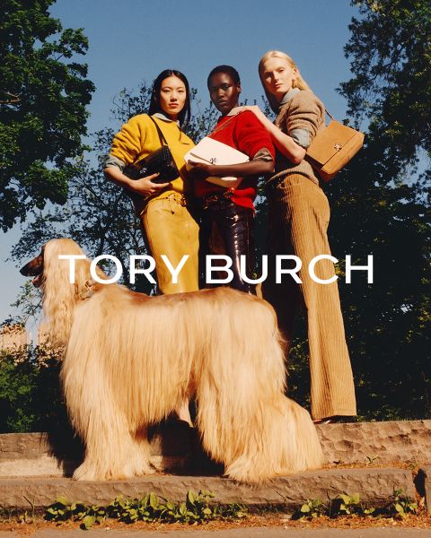 Eine Frau in einem schimmernden Oberteil und einem grünen Rock sitzt neben grünen Geschenkkartons und hält eine geblümte Tory-Burch-Tasche in der Hand; hinter ihr steht ein geschmückter Weihnachtsbaum.