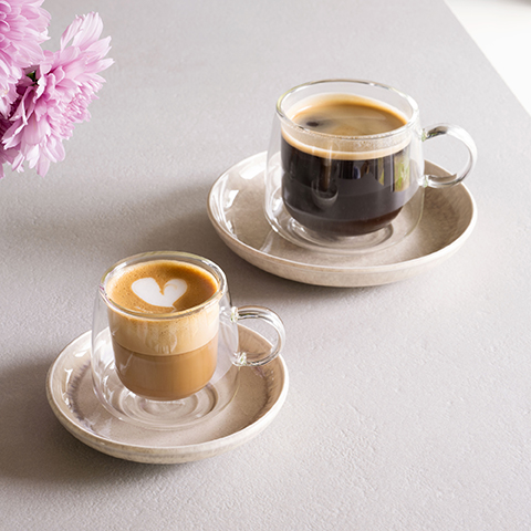 Ein doppelwandiger Glasbecher ist mit cremigem Joghurt gefüllt und mit frischen Himbeeren, Brombeeren und Blaubeeren garniert. Der Becher steht auf einer schwarzen Untertasse mit einem Löffel daneben, vor einem dunklen Hintergrund.