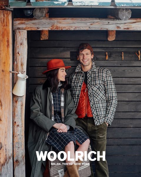 Eine Person, die einen hellbraunen Wintermantel, eine blaue Strickmütze und ein blau kariertes Hemd über einem Kapuzenpullover trägt, steht vor einem einfarbigen Hintergrund. Darunter erscheint das Wort „Woolrich“ zusammen mit den Städten Mailand, Tokio und New York.