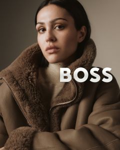 Eine Frau in einem braunen Shearling-Mantel blickt selbstbewusst in die Kamera vor einem neutralen Hintergrund. Das Wort "BOSS" ist in fetten weißen Buchstaben neben ihrer Schulter eingeblendet.