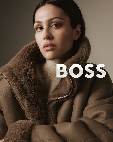 Eine Frau in einem braunen Shearling-Mantel blickt selbstbewusst in die Kamera vor einem neutralen Hintergrund. Das Wort "BOSS" ist in fetten weißen Buchstaben neben ihrer Schulter eingeblendet.
