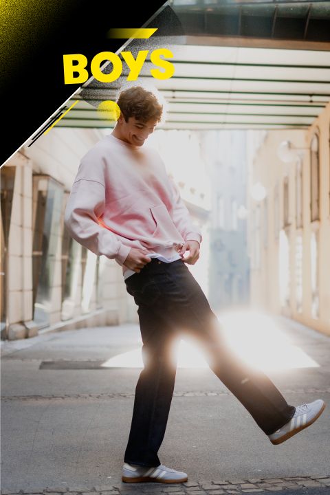Eine Person in einem rosa Sweatshirt, dunklen Jeans und weißen Turnschuhen posiert verspielt in einer sonnenbeschienenen Gasse. Das Wort „BOYS“ ist oben links vor einem Hintergrund mit Farbverlauf deutlich zu sehen.