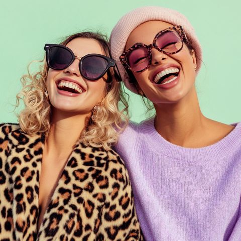 Zwei Frauen lächeln fröhlich und tragen modische Sonnenbrillen. Die eine hat lockiges blondes Haar und einen Mantel mit Leopardenmuster; die andere trägt eine rosa Strickmütze und einen lavendelfarbenen Pullover. Der Hintergrund ist mintgrün.