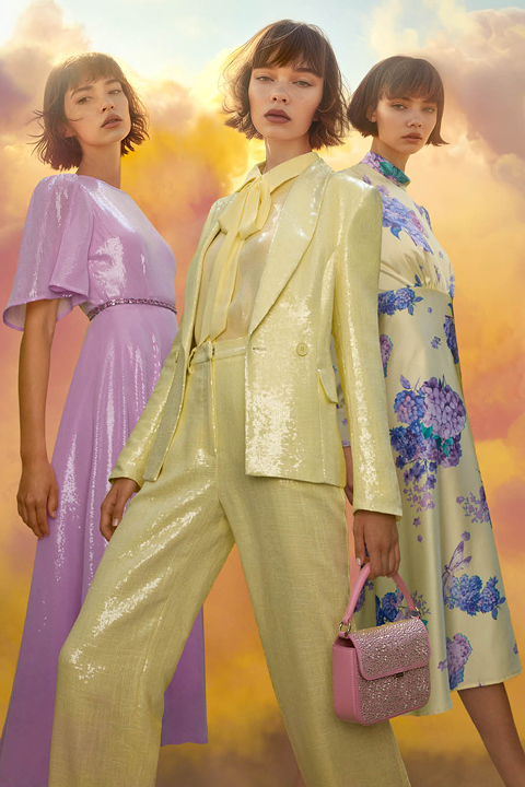 Drei Models posieren vor einem leuchtend bewölkten Himmel. Sie tragen pastellfarbene Outfits: ein lila Kleid, ein gelbes Paillettenkostüm mit Fliege und ein weißes Blumenkleid. Ein Model hält eine pinkfarbene Glitzerhandtasche in der Hand.