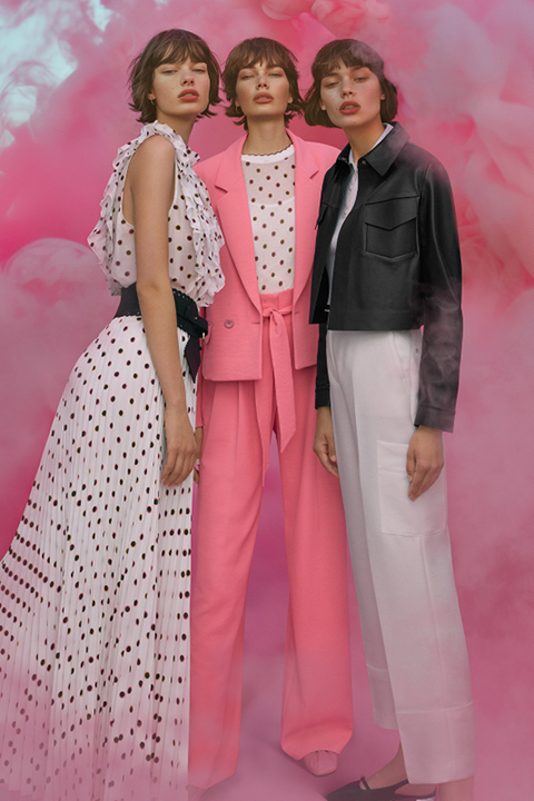 Drei modisch gekleidete Frauen stehen vor einem rosafarbenen, rauchigen Hintergrund. Eine trägt ein gepunktetes Kleid, eine ein rosa Kostüm mit gepunktetem Hemd und die dritte eine schwarze Jacke und weiße Hose. Sie posieren selbstbewusst und strahlen eine moderne, schicke Ausstrahlung aus.