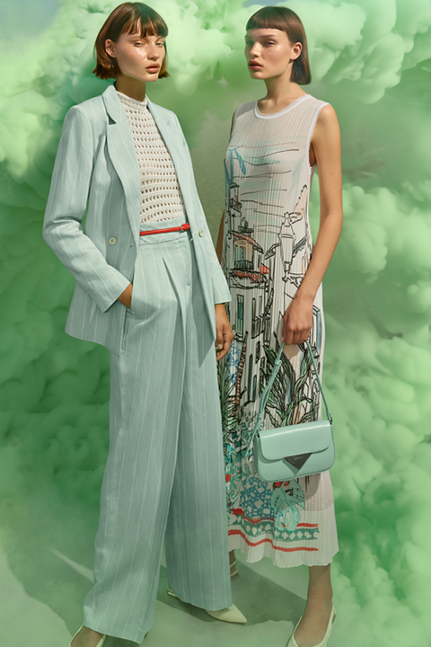 Zwei Models stehen vor einem grünen Rauchhintergrund. Eine trägt einen hellblauen Anzug mit einem weiß gemusterten Oberteil, die andere ein ärmelloses Kleid mit grafischen Mustern. Beide haben kurze dunkle Haare und tragen passende pastellfarbene Accessoires.