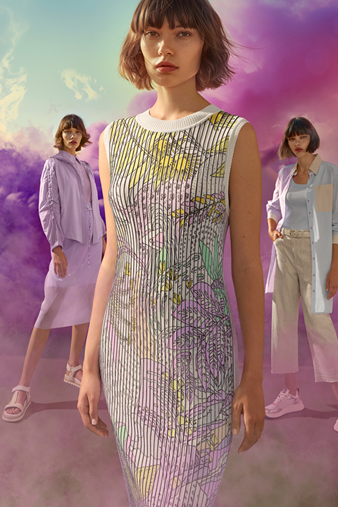 Drei Frauen posieren vor einem leuchtenden Hintergrund aus violetten und rosa Wolken. Die zentrale Figur trägt ein gemustertes Kleid, während die anderen legere, helle Outfits tragen. Die Szene ist farbenfroh und kunstvoll.