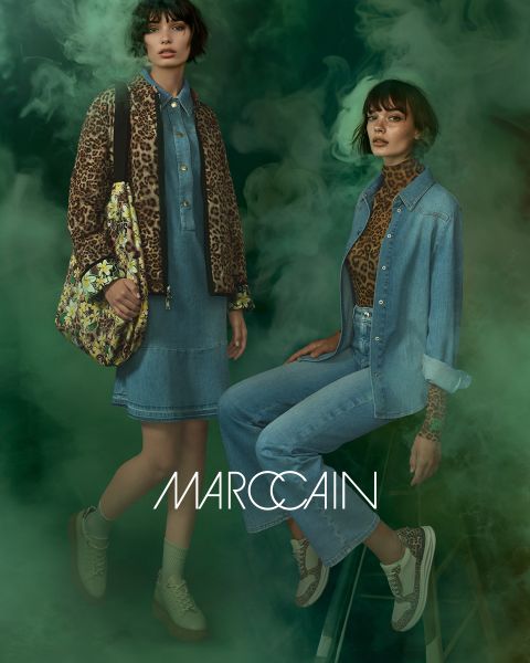 Zwei Models vor einem dunstigen grünen Hintergrund tragen trendige Outfits von Marc Cain. Eine trägt eine Jacke mit Leopardenmuster über einem Jeanskleid und hält eine geblümte Tasche in der Hand. Die andere trägt eine Jeansjacke und -hose sowie ein Oberteil mit Leopardenmuster.