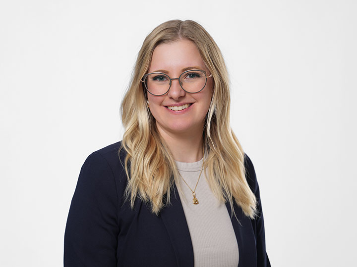 Eine Frau mit langem blondem Haar und Brille lächelt, sie trägt eine helle Jacke und ein gestreiftes Oberteil vor einem schlichten hellen Hintergrund.