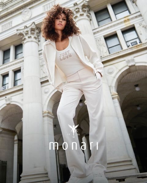 Eine lächelnde Frau, die einen beigen Mantel, ein gestreiftes Hemd, blaue Jeans und einen hellen Schal trägt, steht im Freien vor einem hohen Gebäude. Das Monari-Logo ist am unteren Rand des Bildes zu sehen.