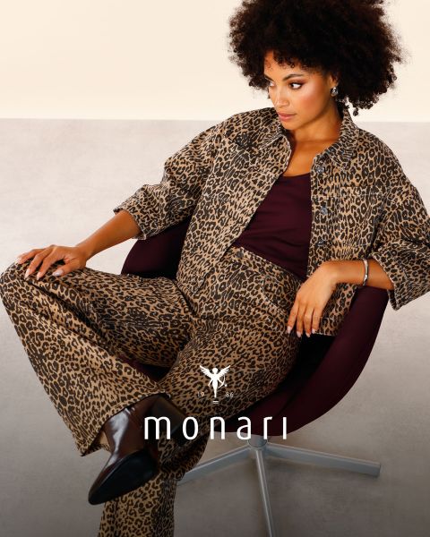 Eine lächelnde Frau, die einen beigen Mantel, ein gestreiftes Hemd, blaue Jeans und einen hellen Schal trägt, steht im Freien vor einem hohen Gebäude. Das Monari-Logo ist am unteren Rand des Bildes zu sehen.