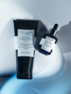 Zwei schwarze Flaschen mit Haar- und Kopfhautpflegeprodukten der Serie „Hair Rituel“ von Sisley vor blauem Hintergrund, eine Tube und eine Tropfflasche.